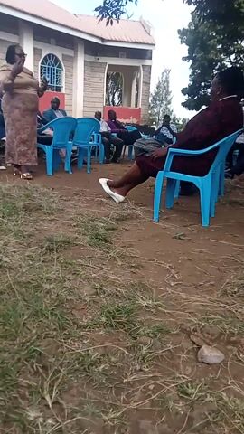 The scene is an outdoor gathering in front of a building. Several people are seated in blue plastic chairs facing the building. A woman in a patterned dress stands in front of the seated group, possibly addressing them. Another woman in a burgundy dress is seen standing partially obscured by a chair. The environment appears to be a yard or garden area with patches of grass and dry ground. The building has a red roof and arched windows. The overall setting seems to be a community event or meeting.