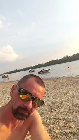 A shirtless man with short dark hair, a goatee, and sunglasses is sitting on a sandy beach. He is looking directly at the camera. The background shows a body of water with several boats on it, and a treeline on the opposite bank. The sky is cloudy and appears to be either late afternoon or early evening. The scene suggests a relaxed, casual atmosphere. There is no visible text in the image.