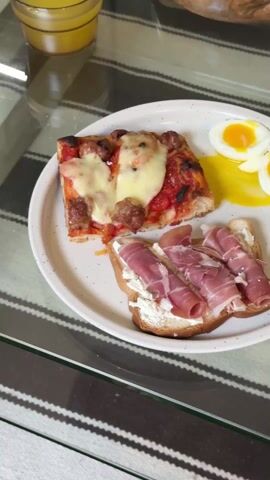 The video shows a plate of food on a glass table. The plate is off-white with brown speckles. The food consists of a square piece of pizza topped with cheese and sausage, a slice of toast topped with white spread and rolled slices of prosciutto, and a halved soft-boiled egg with a runny yolk. The table has a striped design underneath the glass surface. In the background, a jar of amber-colored liquid is partially visible. The camera slowly pans around the plate.