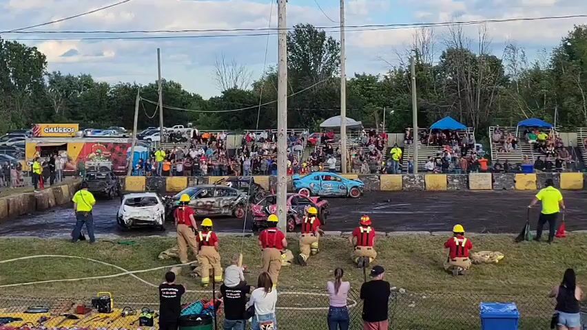 A demolition derby is in progress. A crowd of spectators watches from bleachers. Several wrecked cars are in the arena, some of which appear to be on fire. Firefighters in red shirts and yellow helmets are on the field, some carrying hoses. Several civilians, including children, are also watching from the sidelines. The event appears to be taking place outdoors in the late afternoon or early evening.