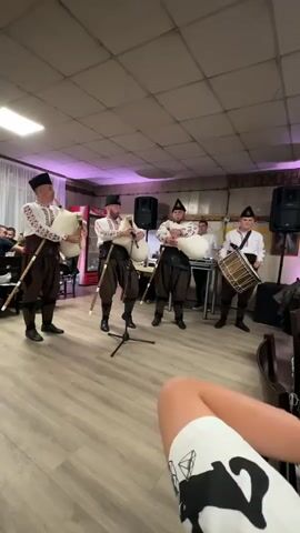 A group of five men are performing music in a room with light-colored flooring and white walls. Four of the men are standing in traditional attire, playing bagpipes. They wear black hats, white long-sleeved shirts with embroidered patterns, and dark baggy trousers. The man on the far right is playing a drum while standing in a white shirt and dark pants. In the background, there is a man in a white shirt operating a keyboard or mixing board. There are several speakers set up, and some people are visible sitting at tables in the left background. The room is dimly lit, with purple uplighting on the walls. The video is shot from a low angle, showing the lower part of a person's legs and feet in the foreground, wearing shorts with a black and white patterned design.