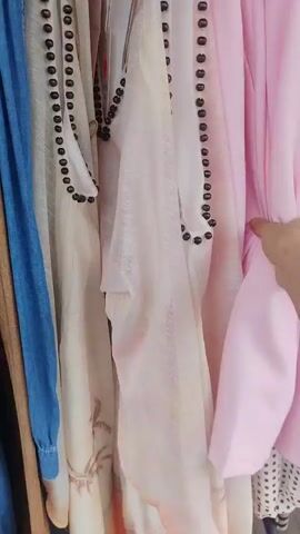 This media file showcases a rack of clothing, primarily in pastel pink and light beige hues, likely in a retail setting. The garments appear to be feminine, with one featuring delicate palm tree patterns. Several pieces are adorned with dark bead detailing along the edges. A hand can be seen adjusting one of the garments, suggesting someone is browsing or trying on clothes. The lighting appears to be natural, possibly daylight, and there are hints of an outdoor environment visible in the background, with a suggestion of a street or sidewalk. The overall impression is one of shopping for fashion, possibly in a boutique or department store. No specific time of day or weather conditions are discernible, beyond the indication of daylight. No legible text is visible in the media file.