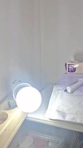 This media file displays a still image, likely captured in a dimly lit room or studio, with a bright light source on the left casting a strong glow. The primary subjects are several objects arranged on a white surface, possibly a shelf or table. In the foreground, a bright, round light bulb is prominently visible, emitting a cool white light. To the right of the light bulb, a white, rectangular package is situated, with some text visible. Beside it, there are two transparent, plastic-encased items that appear to be medical or cosmetic applicators. Further to the right, a small tube of cream or gel is placed, also with some text on its packaging. In the background, a smartphone is propped up, displaying an image of a crowd of people, possibly at an event or concert, wearing colorful clothing. The overall scene appears to be a product display or a staged arrangement, potentially for a review or unboxing video. No specific time of day or weather conditions are discernible, and the emotions conveyed are neutral, focusing on the presentation of the objects. The location context of Kutaglumpang, Indonesia, is provided but not visually evident within the frame.