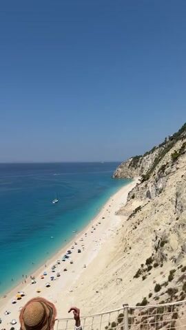The video shows a scenic view of a crowded beach with turquoise waters and a white sand shore. A woman in a bikini and a sun hat is standing on a balcony overlooking the beach. She turns to face the camera and poses. The beach is dotted with colorful umbrellas and people swimming and sunbathing. A sailboat is visible in the distance. The sky is clear blue.