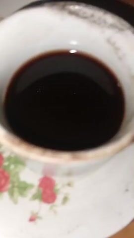 A close-up, slightly shaky video focuses on a white coffee cup filled with dark liquid, resting on a matching saucer. The saucer has a floral pattern with red roses and green leaves. The cup's rim appears a bit worn or chipped. The background is blurry, suggesting a wooden surface. There are no people or explicit actions visible, only the static presentation of the coffee cup and saucer.
