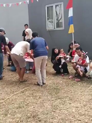 The video shows an outdoor gathering of people, likely a celebration or party, with a grey building in the background. Several people are gathered in a loose circle, with some standing and others kneeling or sitting on the ground. There are colorful flags and banners strung across the scene, suggesting a festive occasion. In the center of the gathering, a few individuals appear to be participating in a game or activity involving plates and food. Some children are present, including a baby being held by an adult. The ground is covered with dry grass or soil, and a yellow rope lies on the ground. The overall atmosphere seems casual and communal.