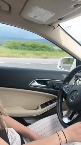 The video shows the interior of a moving car, specifically a Mercedes, from the driver's perspective. The driver, wearing striped pants and sporting white nail polish, is holding the steering wheel. The car's dashboard is visible, featuring a navigation screen displaying a map and music information. Outside the window, the scenery consists of a road, trees, and some buildings under a slightly overcast sky. The car appears to be approaching an intersection.