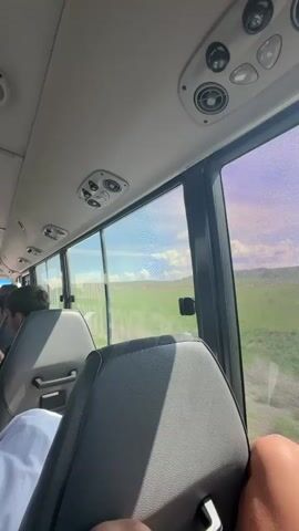 The video shows the interior of a bus moving through a green, grassy landscape. The camera is positioned behind a grey seat, with a person's arm visible. Several rows of empty seats can be seen ahead, and another person’s head is in the left corner. The bus windows show a view of open fields with what looks like a small settlement in the distance. The roof of the bus has overhead compartments and air conditioning vents. The sky is visible through the windows, appearing mostly clear with some cloud cover.