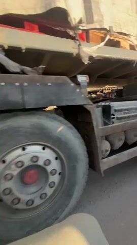 The video shows a close-up, low-angle view of the rear tires and side of a large truck. The truck appears to be moving. The tires are large and show signs of wear and tear. The side of the truck is a dull gray color, possibly dirty or weathered, and there are some red markings on a panel. A blue tarp covers the top portion of the truck bed. The lighting suggests it is daytime, with sunlight casting shadows. The video pans slightly, showing the truck's wheels and undercarriage. The perspective is from someone walking or riding alongside the truck, at a similar height to the lower portion of the vehicle.