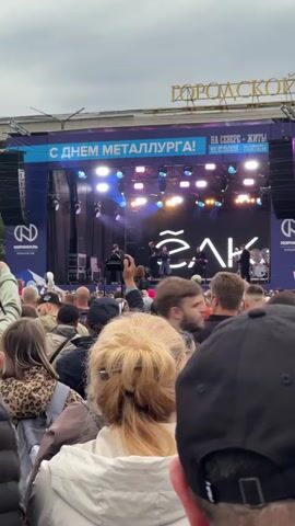 A large crowd of people are gathered outdoors watching a band perform on a stage. The stage is set up with bright lights and a large screen displaying the band's name, which appears to be "ЭЛКА." The band members are visible on stage, playing instruments and singing. A banner above the stage reads "С ДНЕМ МЕТАЛЛУРГА!" which translates to "Happy Metallurgist's Day!". The crowd is diverse in age and includes people of various ethnicities. Many people in the crowd are holding up their phones to record or photograph the performance. The weather appears to be overcast.