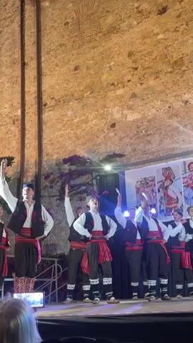 A group of men dressed in traditional folk costumes are performing a dance on a stage in front of a stone wall. The costumes consist of white shirts, black vests with red trim, red sashes around their waists, and dark pants with white stockings. Some of the men wear blue caps. The stage is lit with colorful lights. In the background, there are banners with images of people in similar traditional attire. A person's head is visible in the foreground, possibly holding a phone to record the performance.