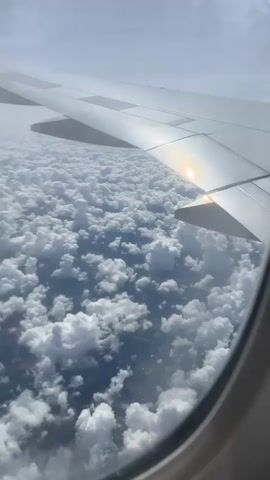 The video captures a view from an airplane window looking down at a vast expanse of white, fluffy clouds. The sun is visible, casting a bright reflection on the airplane's wing, which is prominently featured in the upper right portion of the frame. Below the clouds, glimpses of land can be seen, interspersed with darker patches that suggest shadows or possibly bodies of water. The overall environment is atmospheric and aerial, showcasing a sky filled with cumulus clouds under a hazy sky. The video seems to be a continuous shot, with minimal camera movement, giving a sense of steady flight above the cloud layer.