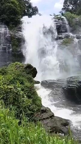 This is a video of a waterfall. The waterfall is large and has multiple tiers. Water is cascading down the rocks. There are trees and vegetation around the waterfall. The sky is visible in the background. The video is in color and appears to be a landscape shot.