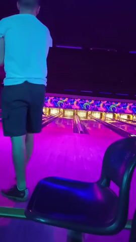 A man in a white t-shirt and black shorts is in a bowling alley. He is holding a bowling ball in his right hand and is about to bowl. The bowling lanes are lit with purple and pink lights, and there are colorful decorations on the walls. A black chair is in the foreground of the image.