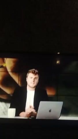 The video shows a TV screen playing a report about a soccer player. The video begins with a man sitting at a table with a laptop and a coffee cup. Two other men stand behind him. The video then cuts to a close up of a yellow soccer jersey with "DORTMUND" and the number "24" on the back. The next shot shows a man in a suit standing in a stadium holding the same jersey. The video then shows the same man wearing the jersey, standing in front of the empty stands of the stadium. The last shot shows the man dribbling a soccer ball through cones on a field.