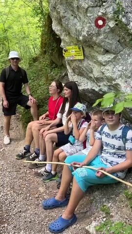 A group of six people are sitting on a rock by a hiking trail, next to a trail marker. A man in a bucket hat and black clothing is standing to their left, leaning against the rock, while five others are seated. A woman is seated in the middle, wearing a red shirt, with two children to her right. A young boy in blue shorts is holding a wooden stick, and to his left is a younger child with his hands over his face. Another woman is seated to the left of the woman in red. The group screams with their hands cupped to their mouths, and the man throws his arms up in excitement. The camera is quickly repositioned to reveal a mountain stream flowing down between rocks. The surrounding environment includes dense foliage, with trees and lush green vegetation.