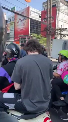 The video shows a young man with brown curly hair sitting on the back of a motorcycle in what appears to be an urban setting. He turns to face the camera, smiling, and holds up a small electronic device, possibly a phone, indicating he is making stickers. There are other people on motorcycles nearby, and a backdrop of buildings and street signs. The overall impression is that he is a tourist or traveler enjoying a moment in a bustling city.