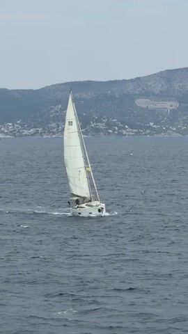 The video shows a white sailboat on a body of water. The sailboat is moving forward, cutting through the water, and its white sail is fully extended. There appear to be people on board. The water is dark blue with some visible waves. In the background, there is a landmass with hills, some buildings, and vegetation. The sky is overcast and grey. At some point, a larger vessel is briefly visible in the far distance.