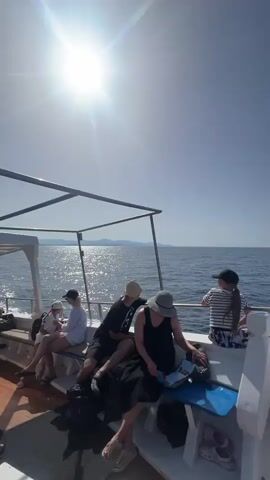 A group of people are on a boat. The sun is shining brightly in the sky, creating a glare on the water. Several individuals are seated on the boat's benches, some looking out at the ocean. One person is wearing a hat and holding a book. Another person is wearing a striped shirt and shorts. The water is blue and calm, with distant land visible on the horizon. The boat appears to be moving.