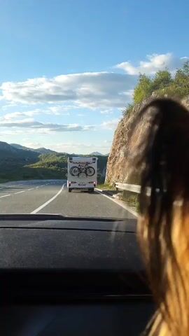 The video shows the view from inside a car driving on a paved highway, following a white camper van. The van has a bike rack on the back with a mountain bike attached. The road has a single white dividing line. The surrounding environment includes green hills and a rocky cliff to the right of the road. The sky is light blue with scattered white clouds. The hair of a person can be seen in the foreground, slightly obscuring the view. The camera car maintains a consistent distance behind the camper van throughout the video. The van's brake lights illuminate.
