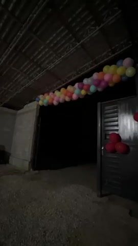 A long garland of multi-colored balloons stretches horizontally across the top of a doorway. The balloons are in shades of pink, yellow, orange, blue, and white. Three red balloons are attached to the right side of the doorway, forming a clover shape. The doorway is dark and appears to be made of metal with horizontal slats. The area is outdoors, with a gravel-like ground surface. Above the doorway, there is a corrugated roof supported by beams. The background is dark, suggesting it is nighttime.