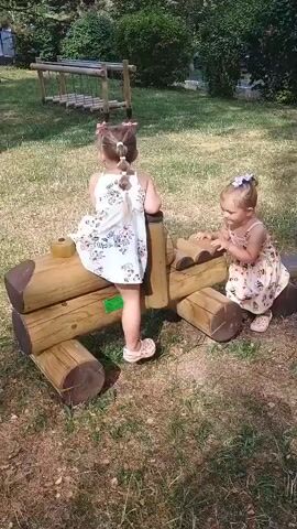 Two young girls are playing on a wooden playground structure resembling a train. The girl on the left, facing away from the camera, is sitting on the structure and wearing a white dress with a floral pattern and pink bows in her hair. The girl on the right, facing the camera, is standing and wearing a patterned dress with bees and purple bows in her hair. The playground is in a grassy area with trees and bushes in the background.
