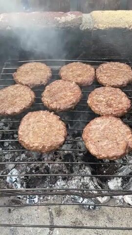 Nine hamburger patties are cooking on a grill. The patties are round and appear to be seasoned. The grill is over hot coals, and smoke is rising from the patties. The grill is situated in an outdoor setting, with brickwork visible in the background. The lighting suggests it is daytime.