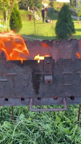 The video shows a close-up of a metal barbecue grill with flames burning inside. The grill is placed on the grass in a yard. In the background, there is a house, trees, and a person walking. The sun appears to be shining, casting a warm light on the scene.