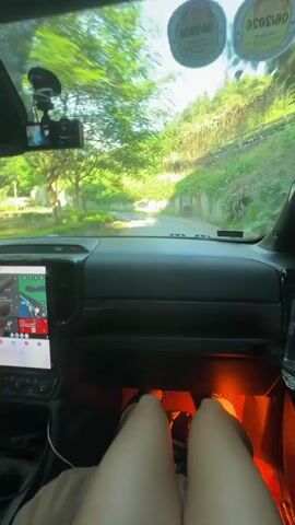 The video is filmed from the perspective of the passenger seat of a car traveling on a narrow road with green vegetation on either side. A person’s legs are visible in the foreground of the frame, positioned above the car’s dashboard and interior lighting. The dashboard features a large touchscreen display and a water bottle in a side compartment. In the final moments of the video, a hand is seen adjusting the vehicle’s visor.
