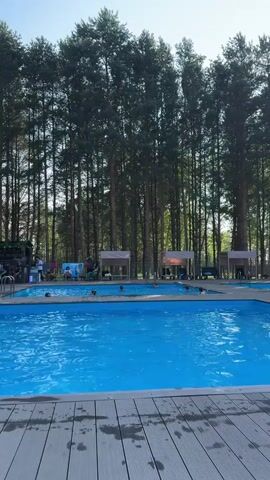 This video captures a bright, sunny day at an outdoor swimming pool complex, with a backdrop of tall, dense pine trees suggesting a wooded or forested location, potentially in Berezovo, Russia. The clear blue water of the swimming pools is the central focus, with several people enjoying a swim. The poolside area features wooden decking and several covered cabanas providing shade.

The activity is clearly recreational swimming. People are visible in the water, some appear to be swimming or wading, while others might be relaxing. On the edge of the pool area, there are also individuals and groups gathered, some sitting, possibly observing or resting. The time of day appears to be daytime, likely late morning or early afternoon, given the bright sunlight and clear sky. The weather seems pleasant and warm, conducive to outdoor swimming.

Notable details include the vibrant blue of the pool water, contrasting with the natural green of the trees and the grey tones of the wooden decking. The sun's glare is visible in the upper right portion of the frame, indicating a strong light source. There are some structures that appear to be changing rooms or shaded relaxation areas, each with a white canopy.

There is some text visible on a green banner in the upper left corner of the video, though it is partially obscured and not clearly legible. The overall mood conveyed is one of relaxation and enjoyment, typical of a summer day at a leisure facility.