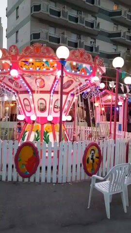 The video shows a carousel ride in motion at what appears to be an amusement park or fair. The carousel is brightly lit with pink and white lights and has decorative elements such as globes and paintings. The ride is enclosed by a white picket fence. In the background, there are buildings and several people, some of whom are on bicycles. The scene is set outdoors, possibly in the evening, given the lighting. There are two white plastic chairs in the foreground.