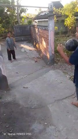 A young boy stands on a concrete path, holding a black ball. He is wearing a dark blue t-shirt and patterned shorts. To his left, a brick wall with peeling paint runs along the edge of the path. In the background, there are trees, houses, and a debris-strewn area. The boy throws the ball towards the wall, which bounces back. He then turns and looks at the ball as it rolls on the ground. The scene is outdoors and appears to be in a rural or semi-urban setting, bathed in sunlight.