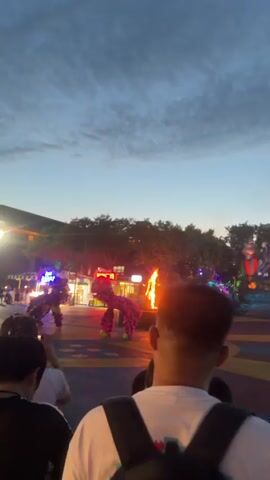 The video begins with a view from behind a crowd of people observing an outdoor event at dusk. In the center of the frame, two performers in elaborate, brightly colored lion costumes are dancing. One lion costume is predominantly pink with black markings, and the other appears to be dark colored. They are performing in front of a large fire pit that is actively burning, casting a warm glow on the surrounding area. The background features a building with illuminated signs, trees, and other people milling about. The sky above is a deep blue with scattered clouds. The camera is positioned at a low angle, looking up towards the performance area. The video continues for a few seconds, showing the continued movement and dance of the lion performers.