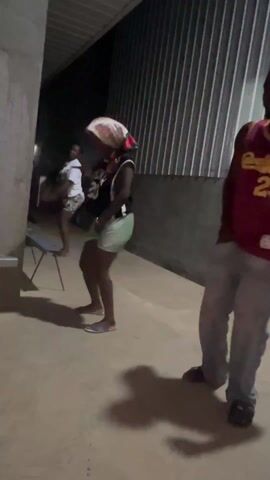 A short video shows three people dancing on a concrete floor in an outdoor area. The location is dimly lit, with a corrugated metal wall visible behind them. The people appear to be of African descent. One person is dancing in a red jersey with the number 23 on it and gray pants. Another person is wearing a dark vest over a black shirt, light green shorts, and a scarf over their head. They are holding something to their mouth, possibly smoking. A third person, partially obscured, is wearing a white top and shorts. A metal folding chair is visible in the background. The three of them appear to be listening to music and enjoying themselves. The shadow of the dancer in the red jersey is prominent on the floor.