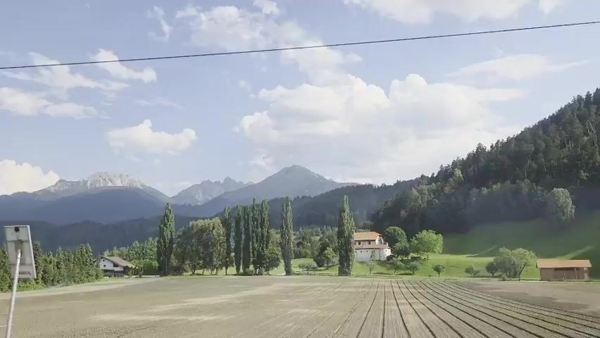 This media file captures a scenic view of a rural Austrian landscape in Unterperfuss. The scene is set during the daytime, under a bright sky with scattered clouds, suggesting pleasant weather. The foreground features a tilled field, indicating agricultural activity.

Prominently visible are several buildings, including a traditional white house with a red roof and a smaller, rustic wooden structure. These buildings are nestled amidst lush greenery and tall, slender trees. In the background, a majestic mountain range rises, with some peaks appearing snow-capped, hinting at a cooler climate or higher altitude. The slopes of the mountains are densely covered with coniferous trees, creating a striking natural backdrop.

The overall atmosphere is one of tranquility and natural beauty. There are no people visible in the frame, emphasizing the peaceful, undisturbed nature of the setting. The composition suggests a journey or a vantage point, perhaps from a moving vehicle, as indicated by the slight movement of the frame and the overhead power line. The clear, bright lighting points to a sunny day, likely during the warmer months given the verdant landscape. No text is visible within the media file.