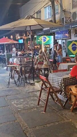 The camera pans slowly to the right across an outdoor seating area of a bar or restaurant. Several small tables covered with black and white checkered tablecloths are set with wooden chairs with red frames. People are sitting at several of the tables, some eating and some on their phones. A couple stands and embraces in front of one table. A man plays a guitar for them. Above the tables are Brazilian flags, posters of football players, and a television. The bar is called "Bar Botafogo" and serves pizza. The ground is uneven and made of concrete tiles. It is night time and lit by street lights.