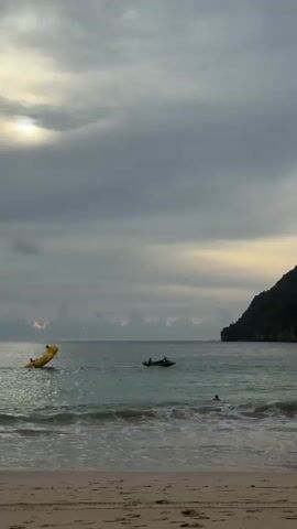 The image captures a serene beach scene at dusk. The sky is overcast with dramatic clouds, allowing a sliver of sunlight to peek through on the left, casting a soft glow on the water. The sea is calm with gentle waves rolling onto the sandy shore.

In the water, several activities are taking place. A bright yellow inflatable banana boat, likely being towed by a hidden boat, is visible on the left side of the frame. Further to the right, a small motorized boat carries a few people. Closer to the shore, a single person is swimming or wading in the water.

The backdrop features a lush, green mountainous island that dominates the right side of the image, its steep slopes disappearing into the cloudy sky. The beach itself is sandy and appears relatively empty, contributing to the peaceful atmosphere. The overall mood is tranquil, suggesting the end of a day at the beach as the light fades. There is no visible text in the image. The location is Meunasahbale, Indonesia.