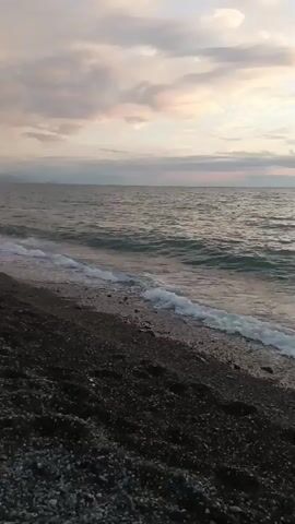 A video captures a pebble beach with waves gently lapping the shore under a cloudy sky. The water is a deep blue-green, and the sky is filled with soft, muted colors. In the distance, there are hints of land and what might be buildings. A few people are visible further down the beach, appearing to be sitting or standing near the water. The overall atmosphere is serene and peaceful.