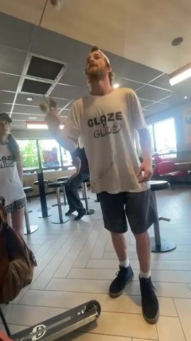 In this video, a man wearing a white t-shirt with "GLAZE 'N GLIDE" printed on it and grey shorts is playing a game of kendama. He is in a casual indoor setting, possibly a fast-food restaurant, with other people in the background and a young girl in a similar shirt to his. The floor is tiled and there are tables and chairs visible. A skateboard is on the floor in the foreground. The man appears focused on the game, and the overall mood seems lighthearted. The lighting suggests it is daytime.