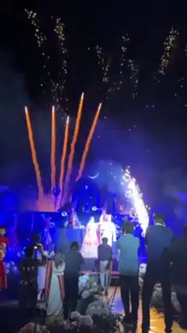 The video shows a wedding taking place at night. In the center of the frame, a bride and groom are standing on a stage, illuminated by bright lights. The bride is wearing a red and white gown, while the groom is in white. Behind them, there's a dark backdrop that appears to be decorated with an Indian deity. Several people are positioned around the stage, including photographers and guests. In front of the couple, sparks are emitting from a machine on either side of the stage, while golden fireworks are shooting up into the night sky. The whole scene is tinged with a blue hue from the stage lighting. The setting suggests a lavish, festive celebration.