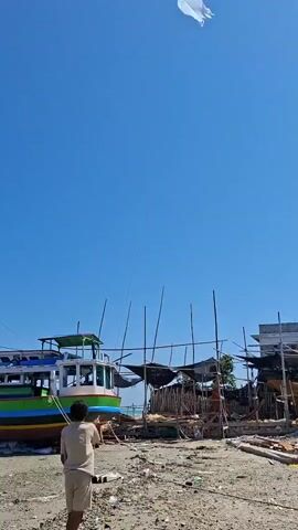 A person stands on a sandy beach, holding a kite string and looking up at a white kite flying in a clear blue sky. In the background, several boats are docked or under repair, with construction materials and debris scattered around. The scene suggests a coastal area, possibly a boatyard or fishing village.