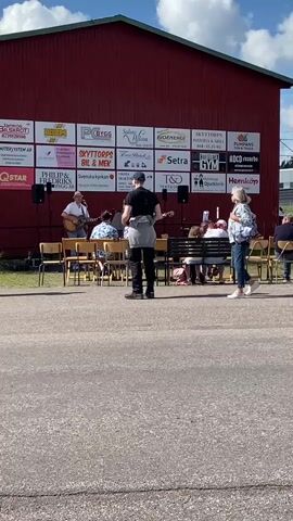 A live musical performance is taking place outdoors in front of a red building. A band of at least two musicians is visible on a makeshift stage. One musician is playing a guitar and appears to be singing, while another musician stands nearby. A small audience is seated on chairs and benches facing the stage. A woman walks across the scene in the foreground, facing the stage and holding a phone. The building behind the stage is adorned with numerous advertisements and logos. The ground in front of the stage is paved, and the surrounding area appears to be grassy with trees in the distance. The sky is blue with some clouds, suggesting fair weather.