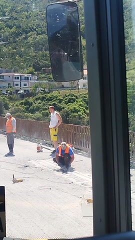 The video captures an outdoor scene, likely on a bridge or elevated road, during a sunny day. The camera appears to be mounted inside a vehicle, looking out a window, with a rearview mirror visible on the left. The perspective is rotated, making the horizon appear vertical.

Initially, two men are visible on a paved surface. One man wears a white t-shirt and yellow shorts, while the other is dressed in an orange safety vest, blue pants, and a hard hat. Construction barriers with red and white stripes and a yellow flashing beacon are positioned on the road. A yellow cable or hose stretches across the ground.

As the video progresses, a third man in a light blue shirt walks past. A yellow compact utility vehicle, resembling a mini excavator or loader, slowly backs up, navigating around the construction barriers. The background features a verdant landscape of rolling hills covered in dense green trees, with several white residential buildings interspersed among the foliage.

A dark sign with white text is prominently displayed, showing an arrow pointing right and the words "EJADIN tours" and "ULCINJ - PODGORICA," indicating destinations in Montenegro. The scene suggests ongoing road construction or maintenance work under clear skies in a hilly, possibly coastal, region.
