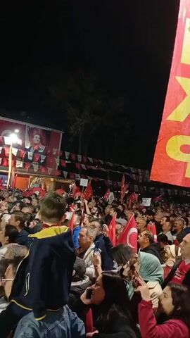 Here is a description of the video:

The video shows a large crowd of people gathered outside at night. Many of them are holding Turkish flags. The crowd is facing a building decorated with images of a man, possibly a political figure. Red banners with yellow lettering are held high in the air. Some individuals are holding up their phones, possibly taking pictures or videos. A small child is perched on someone's shoulders, looking toward the stage. The scene suggests a political rally or public gathering.