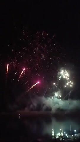 The video shows a nighttime fireworks display over a body of water. Multiple fireworks are exploding in the dark sky, creating colorful bursts of light and trails of smoke. The reflections of the fireworks are visible on the surface of the water. In the foreground, the silhouettes of people can be seen watching the display.