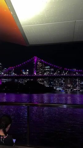 A view of a bridge illuminated with purple lights at night is captured. The bridge spans across a body of water, which reflects the purple light. In the background, a city skyline with numerous lit buildings is visible. The video appears to be filmed from an elevated position, possibly from a balcony or a boat, as a railing is visible in the foreground, and a ceiling with light fixtures is overhead. The water has ripples, indicating movement. The overall scene is a nighttime cityscape with a prominent, colorful bridge as the focal point.