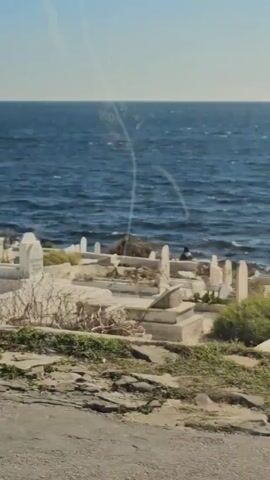 The video shows a view of the ocean from a moving vehicle. In the foreground is a cemetery with white tombs. A palm tree is partially visible in the frame. The ocean is blue with small waves. The sky is clear and blue. There are some rocks and vegetation near the shore. The view is seen through a window, as evidenced by reflections on the glass.