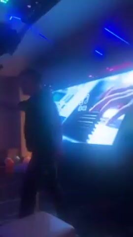 The video reveals the interior of a karaoke room with a lively atmosphere. Initially, the camera focuses on a large screen displaying hands playing a keyboard, illuminated in bright blue. The camera then pans across the room, showcasing the pink and purple neon lighting, decorative balloons scattered on the floor, and round windows lining the wall. A man walks into the frame, briefly obscuring a bright orange storage container. The camera angle then shifts upward, emphasizing the setup of snacks on a table, before the video concludes.