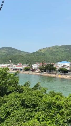 The video shows a view of a small coastal town from a balcony. In the background, there are lush green hills. The town consists of various buildings, mostly white, with a few having red or blue roofs. There is a line of boats pulled up on the shore, and a body of water separates the viewer from the town. The foreground is dominated by green bushes. The sky is clear and blue. The camera slowly pans to the right.
