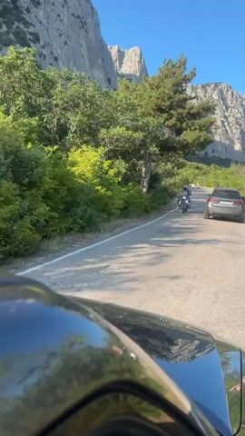 This video shows a perspective from inside a vehicle driving on a paved road. The road is bordered by lush green trees and vegetation on the left side, with dramatic, rocky cliffs in the background. A motorcycle with a rider wearing a helmet and light-colored jacket is approaching and passes the vehicle. Further down the road, another dark-colored SUV is visible driving in the same direction. The sky is clear and blue. The camera is pointed forward, capturing the scene through the vehicle's windshield.